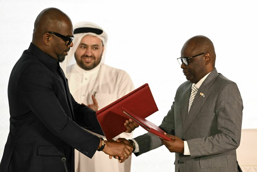 Zwei Männer im Anzug übergeben sich rote Dokumentmappen. Eine Person im Hintergrund klatscht anerkennend