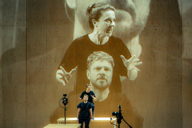 Zwei Schauspieler, live von einer Kamera aufgenommen. Ihr Bild wird überlebensgroß hinter sie auf eine Wand projiziert
