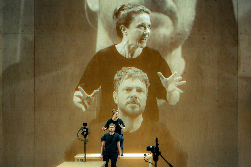 Zwei Schauspieler, live von einer Kamera aufgenommen. Ihr Bild wird überlebensgroß hinter sie auf eine Wand projiziert