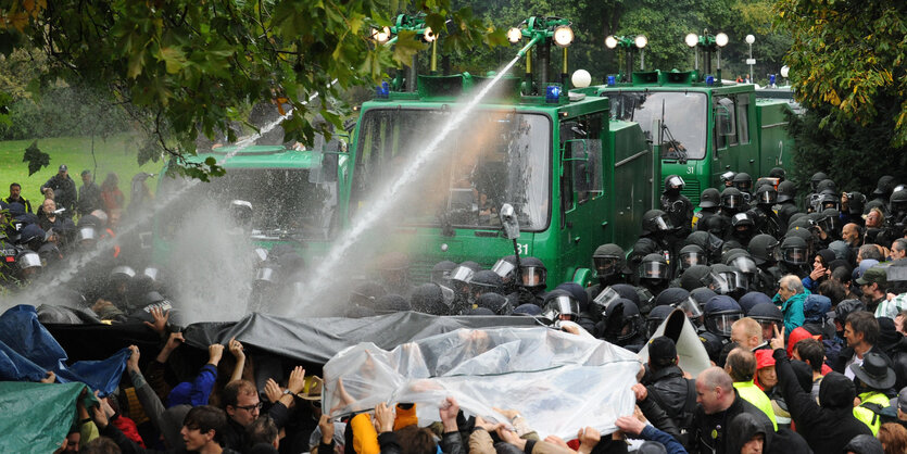 Wasserwerfer zielen auf DemonstrantInnen, die versuchen, sich mit Planen zu schützen