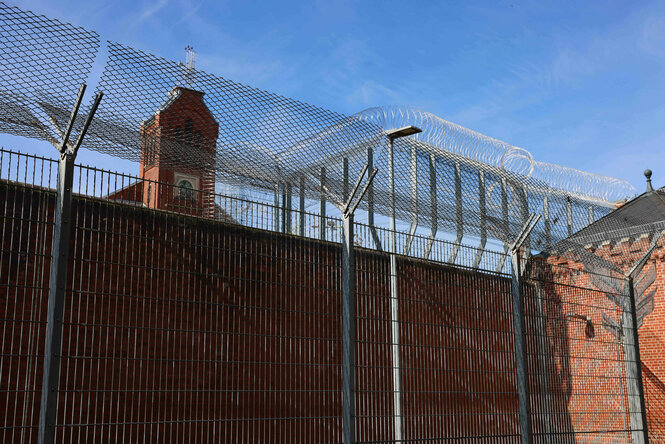 Ein gesicherter Gefängnishof mit rotem Backsteinmauerwerk, Stacheldraht und einem alten Kirchturm im Hintergrund unter blauem Himmel