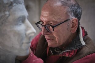 Bildhauer Joachim Sauter, rote Jacke und braune Weste, legt mit konzentriertem Blick durch seine Brille Hand an die graue Skulptur von Betty Rosenfeld, die am linken Bildrand im Profil zu sehen ist.