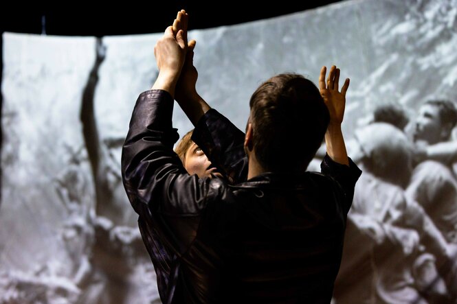 Ein Mann und eine Frau stehen sich gegenüber und berühren sich an der Hand. Im Hintergrund schwarz-weiße Bilder