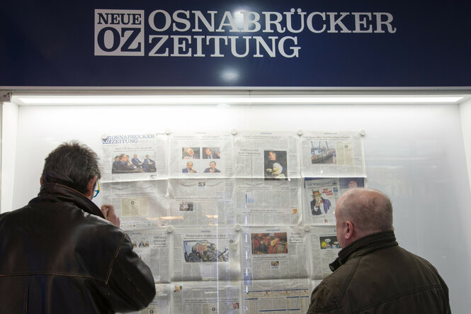 Zwei Männer stehen nachts vor einem beleuchteten Schaukasten der „Neuen Osnabrücker Zeitung“ und lesen die ausgehängten Zeitungsseiten