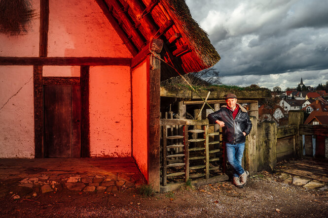 Karl Banghart steht neben einem rot beleuchteten Fachwerkhaus, er lehnt am Zaun eines Stalles, im Hintergrund ist die Stadt Oerlinghausen zu sehen