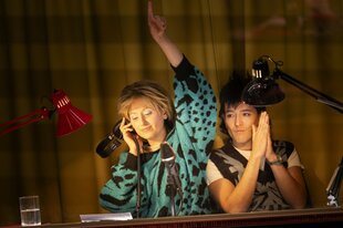 Eine Darstellerin, ein Darsteller, gekleidet in Spät-DDR-Chic, sitzen in einem von Schreibtischlampen erhellten Pult mit Mikrofon, die Frau hebt den linken Arm und hält mit der rechten ihren Kopfhörer