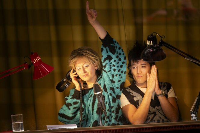 Eine Darstellerin, ein Darsteller, gekleidet in Spät-DDR-Chic, sitzen in einem von Schreibtischlampen erhellten Pult mit Mikrofon, die Frau hebt den linken Arm und hält mit der rechten ihren Kopfhörer