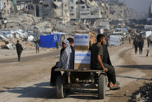 Menschen fahren auf einem beladenen Wagen durch eine Trümmerlandschaft.