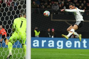 Ein Fußballspieler erzielt im Sprung einen Treffer. Zu sehen ist auch der gegnerische Torhüter