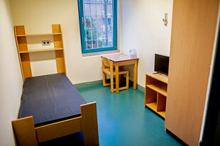 In einer Gefängniszelle stehen ein Bett mit dunkler Matratze, Holzregal, kleiner Tisch mit Stuhl, Fernseher, Wasserkocher, vergittertes Fenster, türkiser Boden.