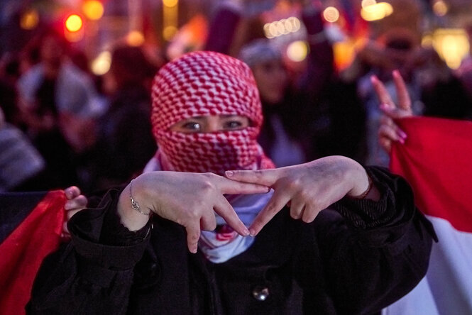 Eine vermummte Person zeigt mit den Fingern ein Dreieck auf einer Demonstration