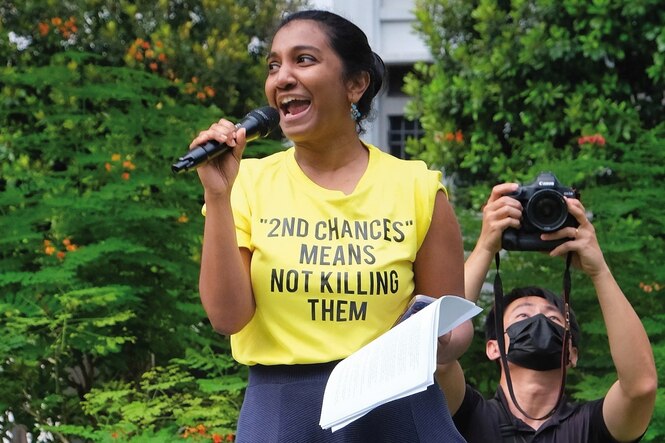 Eine junge Frau in gelbem T-shirt mit aufgedruckter Schrift spricht in ein Mikrofon
