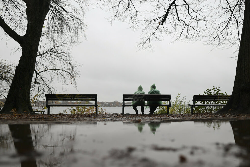 Zwei Menschen mit grünen Regenponchos sitzen bei leichtem Dauerregen auf einer Bank an der Außenalster.