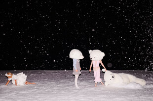 Über eine verschneite Fläche vor einem schwarzen Hintergrund kriecht eine junge Frau nach links, zwei andere, mit Bärenmasken, unterhalten sich