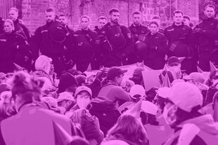 Eine große Gruppe von demonstrierenden Menschen sitzt dicht gedrängt auf einer Straße. Hinter ihnen steht eine Reihe von Polizist*innen in Schutzkleidung und mit Helmen, die die Situation beobachten.