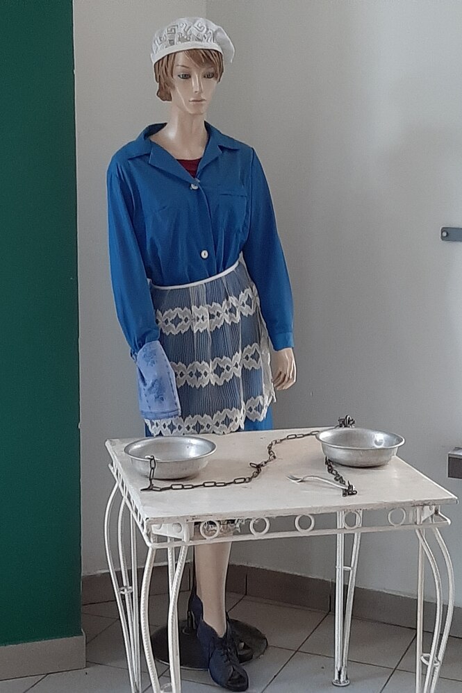 Eine Puppe mit weißer Haube auf dem Kopf steht an einem Tisch, auf dem zwei Blechnäpfe stehen