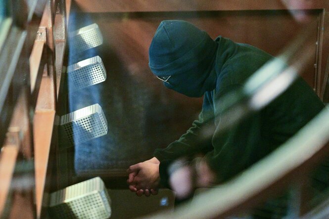 Eine vermummte Person sitzt auf einer Anklagebank