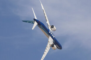 Eine Boeing 777X mit weißen Tragflächen und blauem Rumpf fliegt nach rechts unten geneigt. Der Himmel ist blau mit dünnen Wolken.