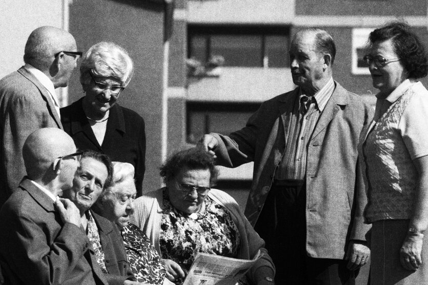 Eine Gruppe von Rentner steht in einem Hof, im Hintergrund sind Häsuer mit Balkonen zu sehen. Eine sitzende Frau liest in einer Zeitung, die anderen Personen sind im Gespräch