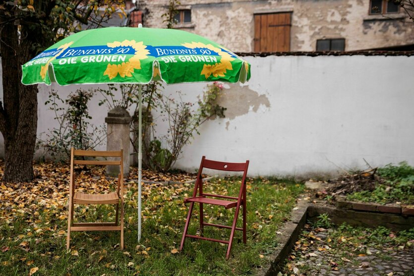 Herbstliche Stimmung in einem Hinterhof,ein Sonnenscirm mit demLogo der Partei Die Grünen schützt zwei Stühle, die auf mit Laub bedecktem Rasen stehen