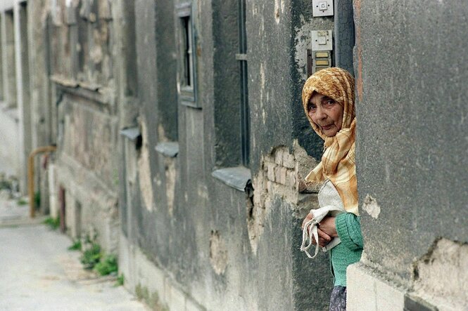 Eine alte Frau mit Kopftuch lugt vorsichtig hinter einem Mauervorsprung
