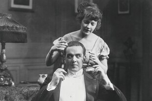 Schwarz weiss Foto aus den 20er Jahren - eine junge Frau steht in einem bürgerlichen Wohnzimmer hinter ihrem gut gekleideten Mann und und versucht mit ihren Händen seinen Kopf zu umschliessen