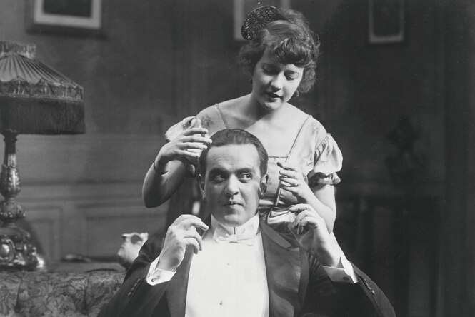 Schwarz weiss Foto aus den 20er Jahren - eine junge Frau steht in einem bürgerlichen Wohnzimmer hinter ihrem gut gekleideten Mann und und versucht mit ihren Händen seinen Kopf zu umschliessen