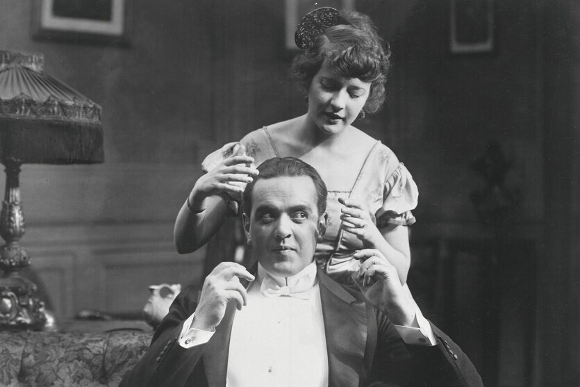 Schwarz weiss Foto aus den 20er Jahren - eine junge Frau steht in einem bürgerlichen Wohnzimmer hinter ihrem gut gekleideten Mann und und versucht mit ihren Händen seinen Kopf zu umschliessen