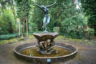 Ein Brunnen steht in der Parkanlage, in der Mitte des Brunnens steht auf einem Sockel der von Menschen in sitzender Haltung getragen wird. Oben eine Bronzestatue, ie eine nackte junge Tänzerin mit ausgebreiteten Armen zeigt.