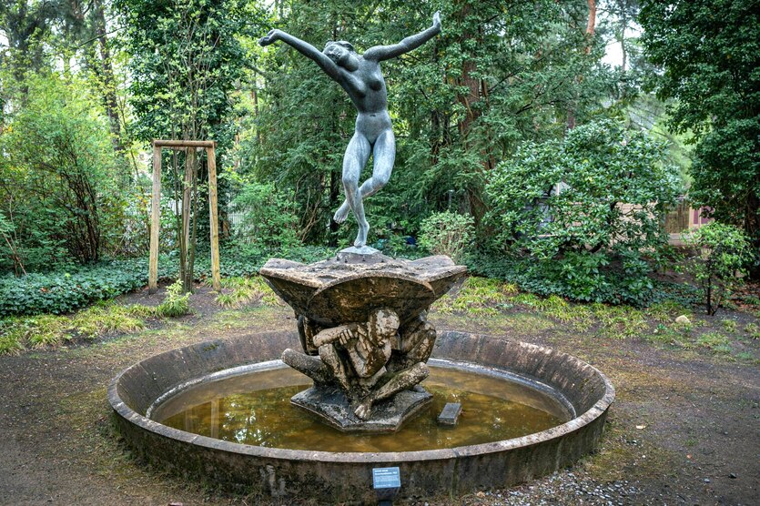 Ein Brunnen steht in der Parkanlage, in der Mitte des Brunnens steht auf einem Sockel der von Menschen in sitzender Haltung getragen wird. Oben eine Bronzestatue, ie eine nackte junge Tänzerin mit ausgebreiteten Armen zeigt.