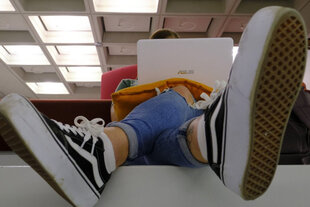 Ein Mensch sitzt vor aufgeklapptem Laptop in einem Schulzimmer und überschlägt die Beine, im Vordergrund die Schuhsohlen seiner Vans-Sneakers.