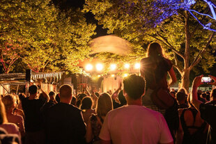 Festliche Lichtstimmung an einer Bühne unter dem Laubdach zweier Bäume, Menschen schauen und klatschen in Richtung der Vorstellung.