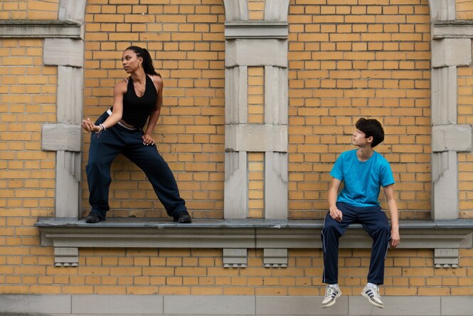 Eine Tänzerin steht auf einem Sims einer Fassade aus Backsteinen, neben ihr sitzt ein Junge in einer Nische