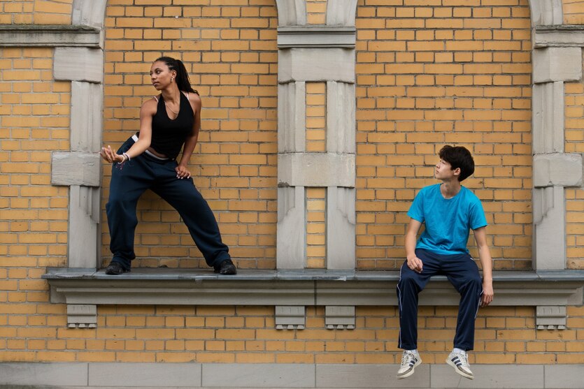 Eine Tänzerin steht auf einem Sims einer Fassade aus Backsteinen, neben ihr sitzt ein Junge in einer Nische