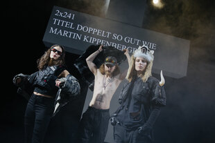 Drei Menschen in schwarzer Kleidung vor einem großen Kreuz