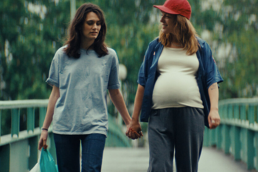 Zwei Frauen halten Händchen und sehen glücklich aus. Die rechte Frau ist schwanger.