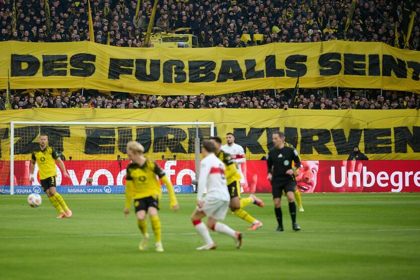Im Hintergrund große Transparente, auf denen zu lesen ist: Soll das die zukunft des Fußballs sein". Davor Spieler des BVB und des VfB Stuttgart