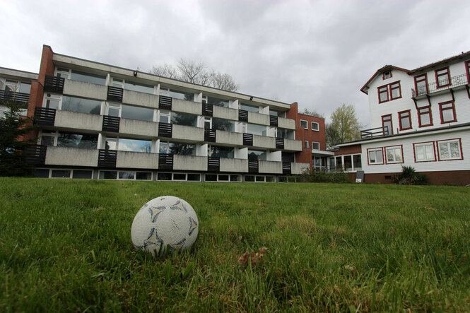 Ein Fußball liegt auf einer grünen Wiese vor einem modernen Mehrfamilienhaus mit Beton- und Ziegel-Elementen unter bewölktem Himmel.