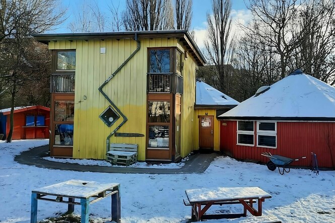 Ein leerer verschneiter Platz mit gelbem Holzhaus mit großen Fenstern und Zickzack-Rinne und roter Jurte