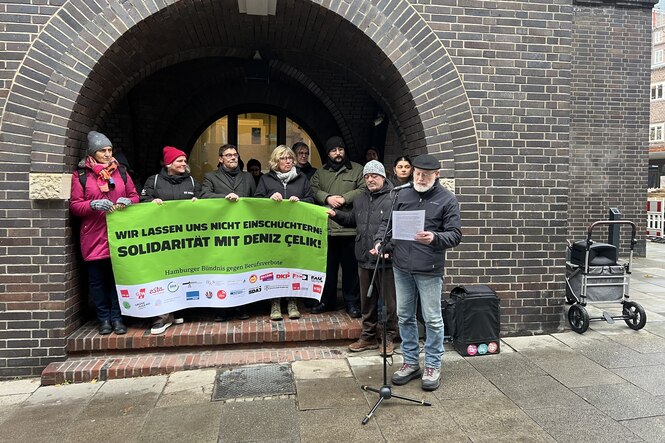 Eine Gruppe Demonstrierender vor einem Backstein-Torbogen hält ein grünes Banner: „Wir lassen uns nicht einschüchtern! Solidarität mit Deniz Çelik!“ Ein Redner spricht ins Mikrofon.