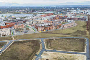 Luftaufnahme eines modernen Wohnviertels mit roten und beigen Mehrfamilienhäusern, teils fertig, teils im Bau, umgeben von Feldern.