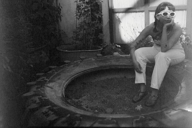 Peki Momés sitzt in einem alten LKW-Reifen und lehnt an einer Hauswand. Sie trägt eine schicke weiße Sonnenbrille