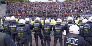 Eine voll besetzte Kurve in einem Fußballstadion, davor Polizisten