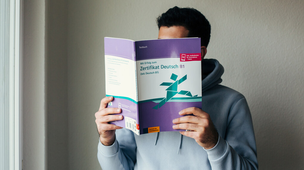 Eine Person hält sich ein Deutschkursbuch vor das Gesicht.