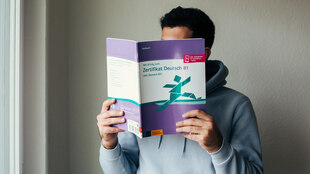 Eine Person hält sich ein Deutschkursbuch vor das Gesicht.