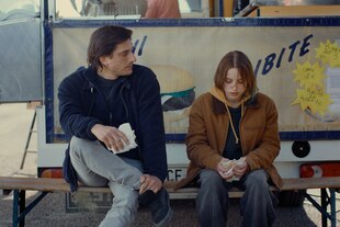 Luca Marinelli und Juli Grabenhenrich sitzen vor einem Imbiss auf einer Bierbank.