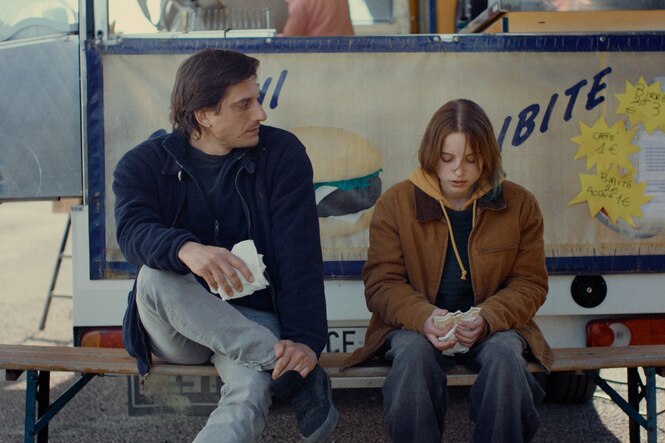 Luca Marinelli und Juli Grabenhenrich sitzen vor einem Imbiss auf einer Bierbank.