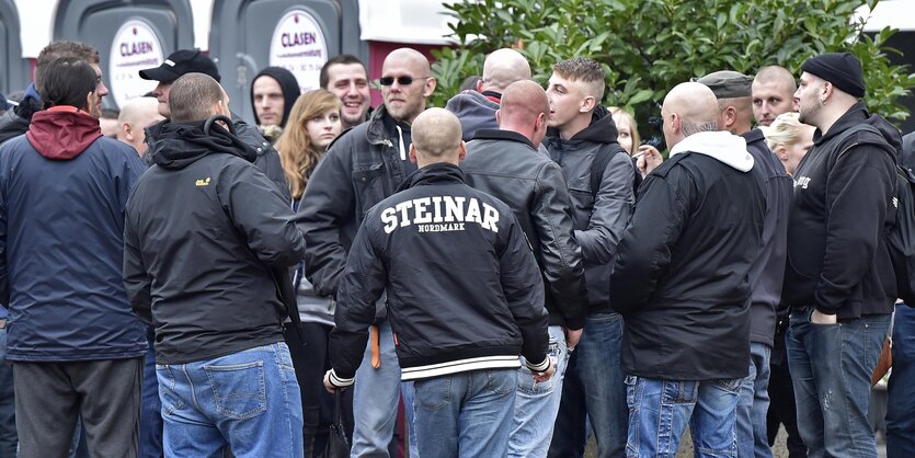 Eine Gruppe Nazis mit Glatzen steht in einer Gruppe und tut, was sie immer tun: Blödsinn reden und denken. In der Bildmitte steht einer mit einer Thor Steinar Jacke.