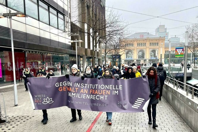 Menschen demonstrieren vor Hannovers Hauptbahnhof mit einem violetten Banner, auf dem steht: "Gegen instittutionelle Gewalt im Familienrecht"