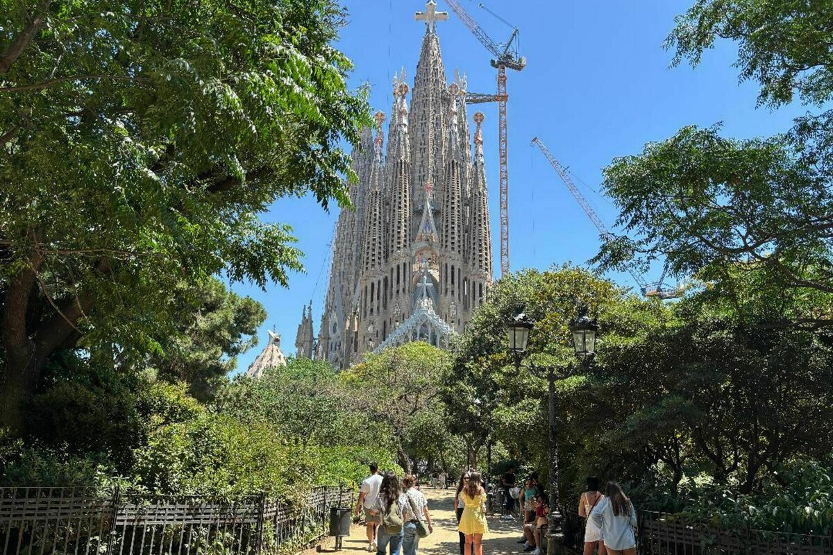 Touristen suchen in einem Park in Barcelona nach einem schattigen Plätzchen. Im Hintergrund ragt die berühmte Sagrada Familia empor.